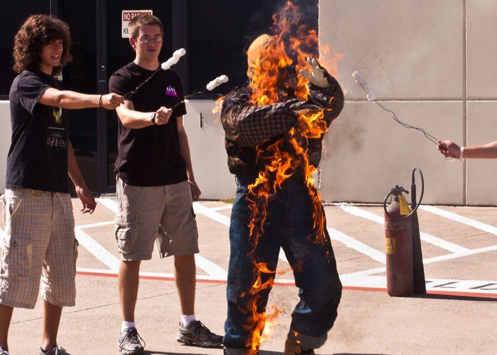 Two people hold sticks with marshmallows over a person wearing a fire-resistant suit fully engulfed in flames.