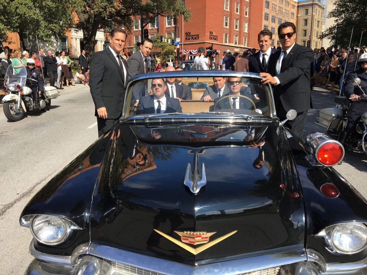 Men in suits stand around and sit inside a vintage black convertible car on a sunny street.