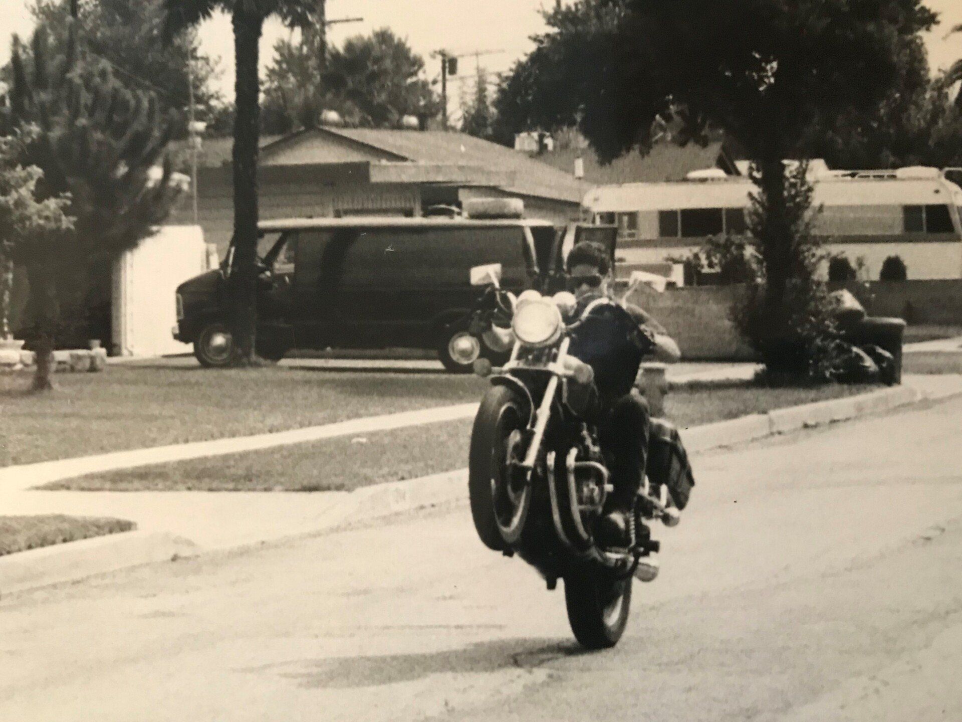A motorcyclist performs a wheelie on a residential street in front of a parked van and a trailer.