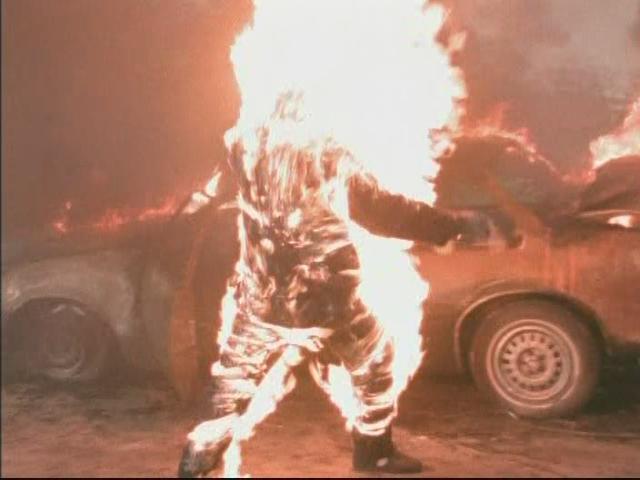 A person fully engulfed in flames stands before a burning car in a dark, fiery scene.