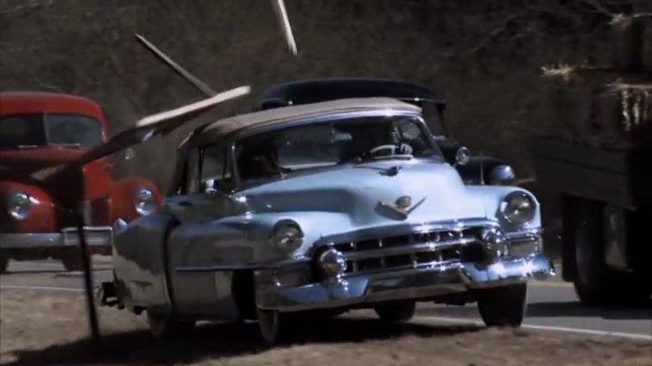 A light blue vintage Cadillac convertible driving on a road near a red truck and a truck carrying hay.