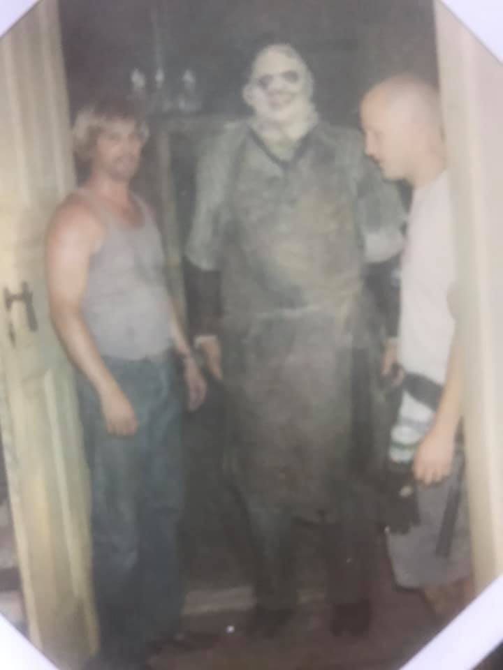 A man in a sleeveless shirt and a man in a t-shirt stand beside a person dressed as Leatherface in a dark room.