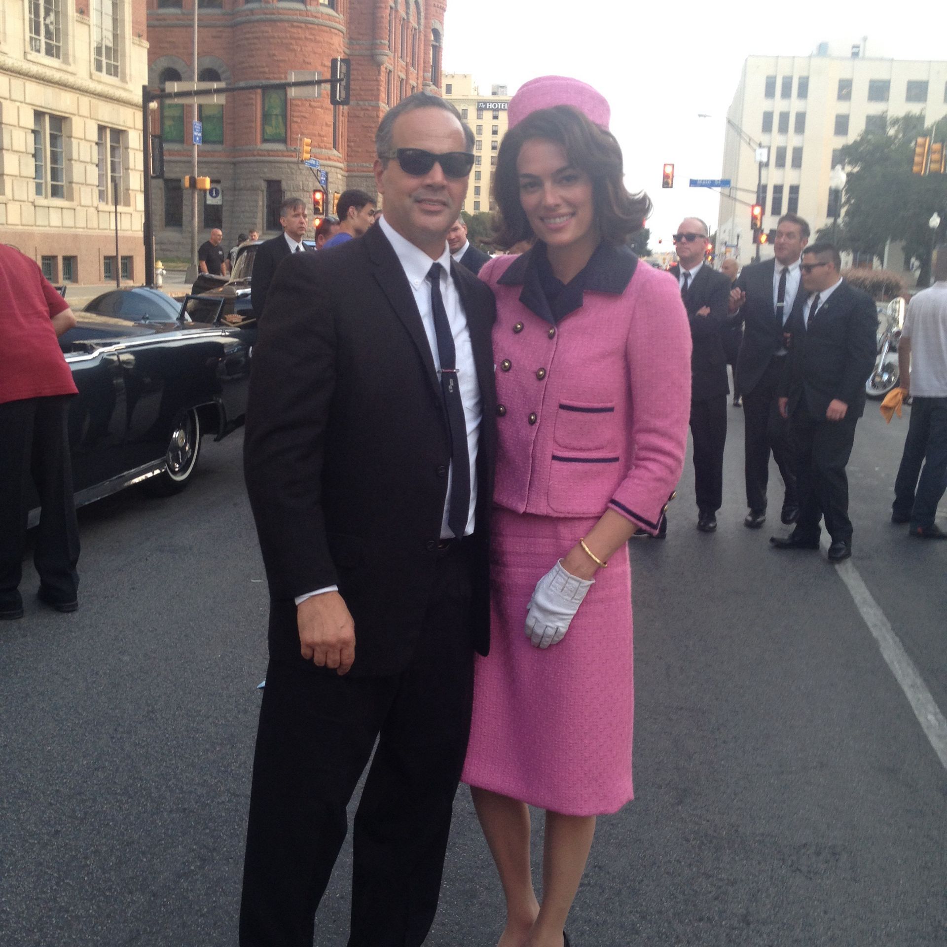 A couple in 1960s-style formal wear, including a pink suit and pillbox hat, poses on a street with onlookers and cars.