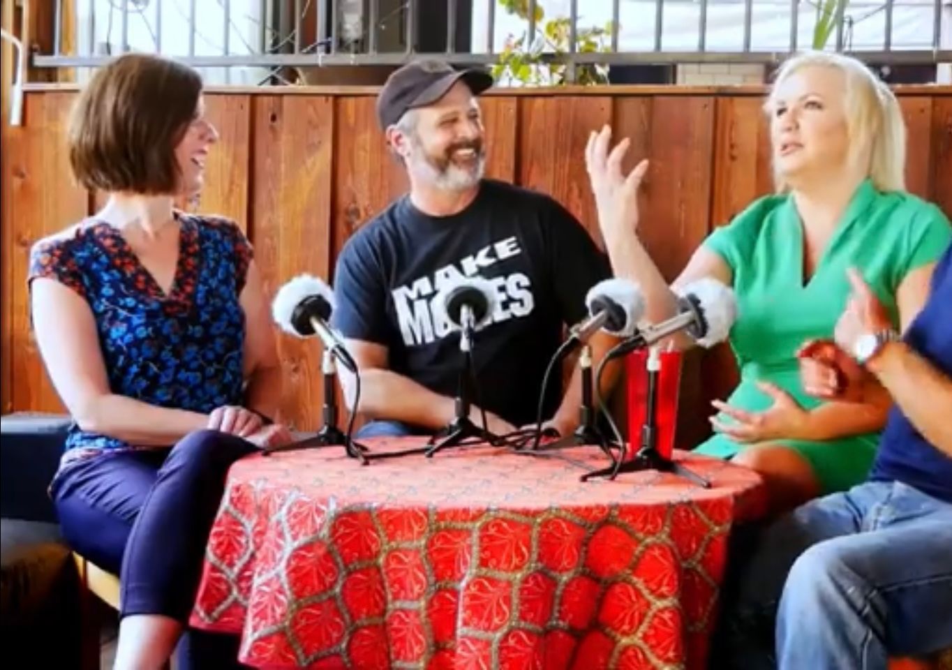 Four people sit around a table with microphones, appearing to record a podcast in a room with wood-paneled walls.