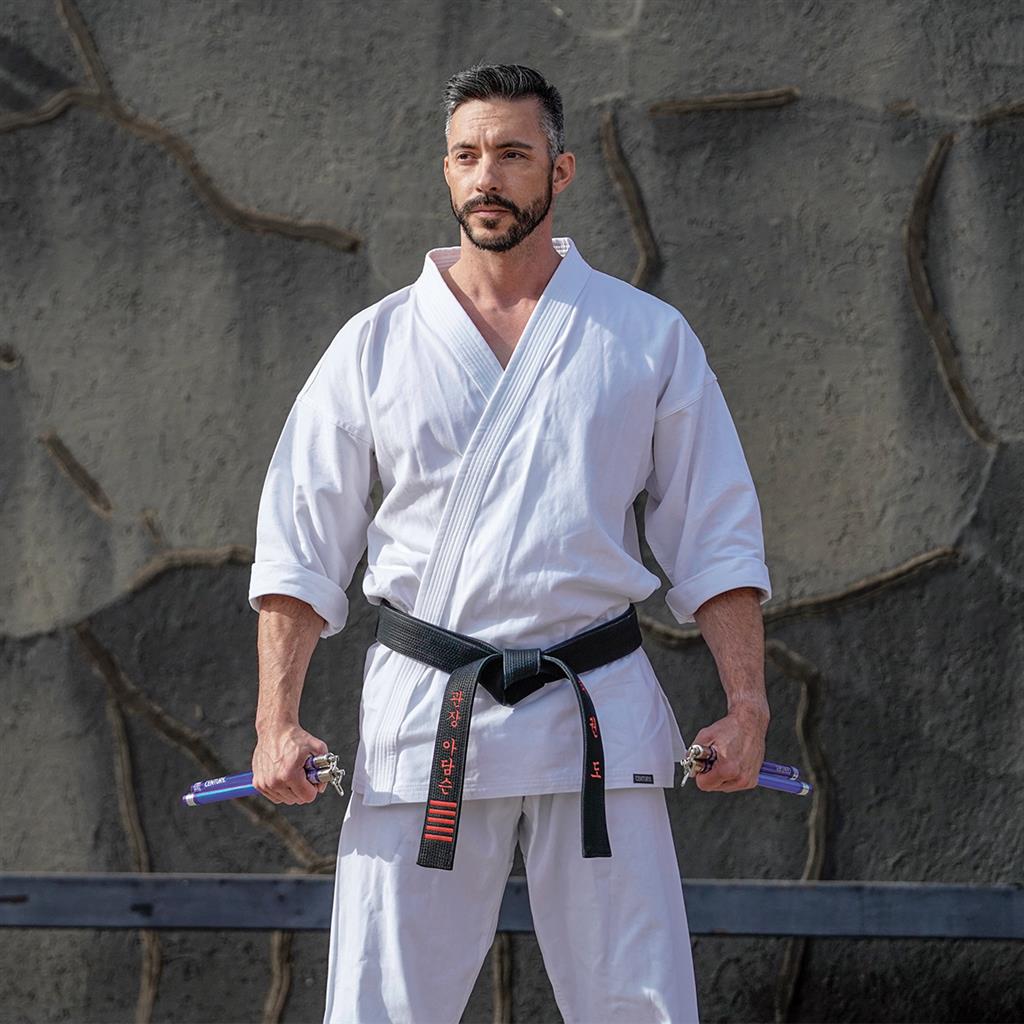 A martial artist wearing a white gi and black belt holds nunchucks against a textured, stone-like wall.