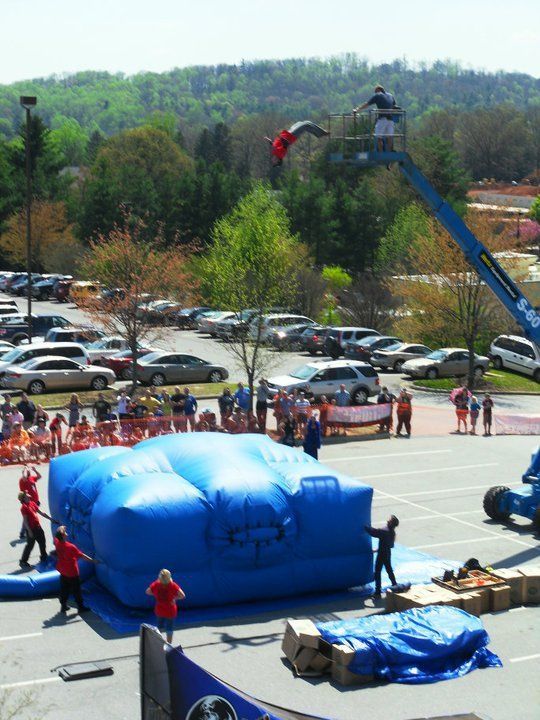 A person jumps from a tall mechanical lift onto a large blue inflatable stunt bag in a parking lot with spectators nearby.