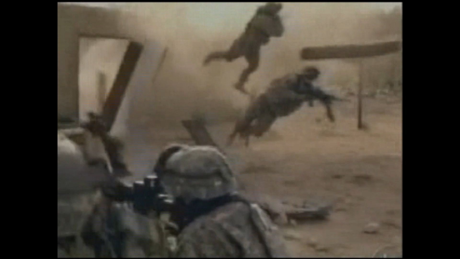Soldiers in camouflage gear leap through the air away from a dusty, debris-filled explosion near a building.