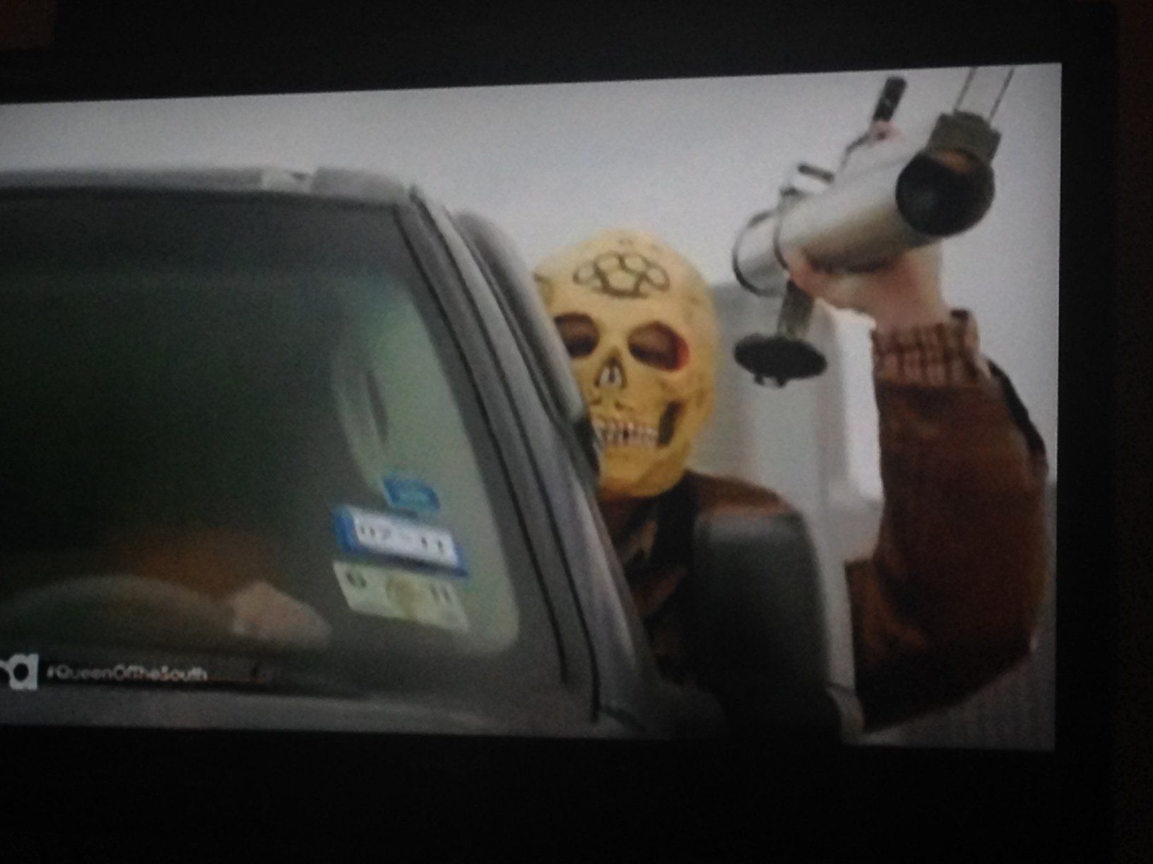 A person wearing a skull mask holds a rocket launcher out the window of a car.