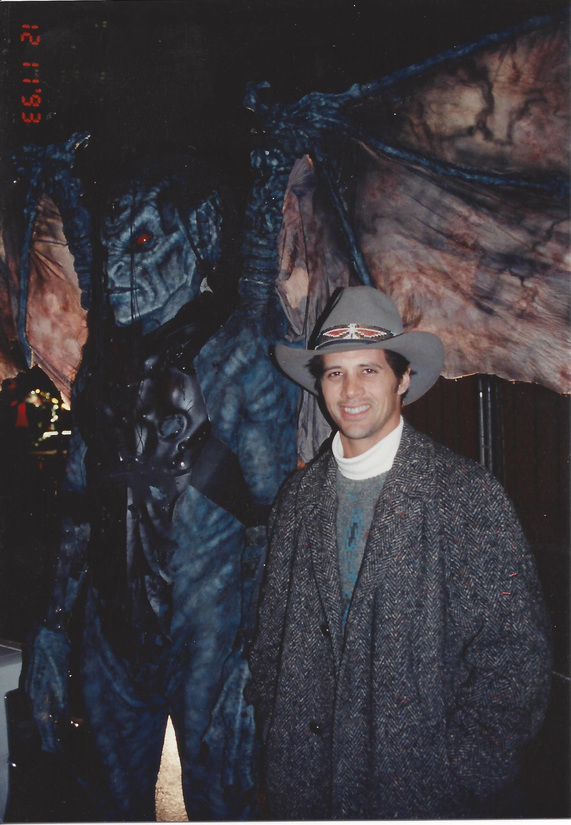 A person in a gray cowboy hat and coat stands smiling next to a tall, dark, winged demon costume with red eyes.