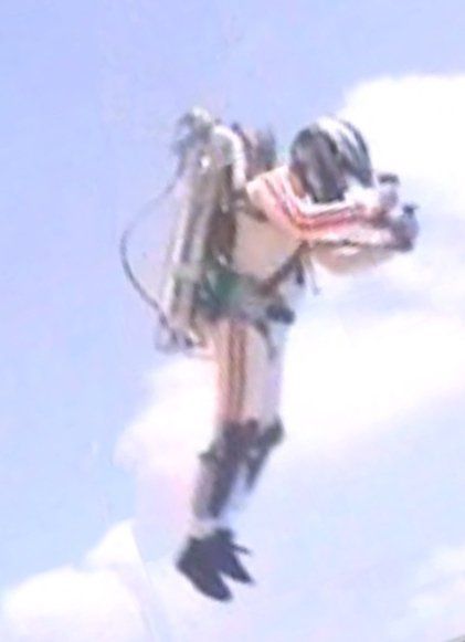 A person wearing a flight suit and helmet is suspended in mid-air with a jetpack strapped to their back.