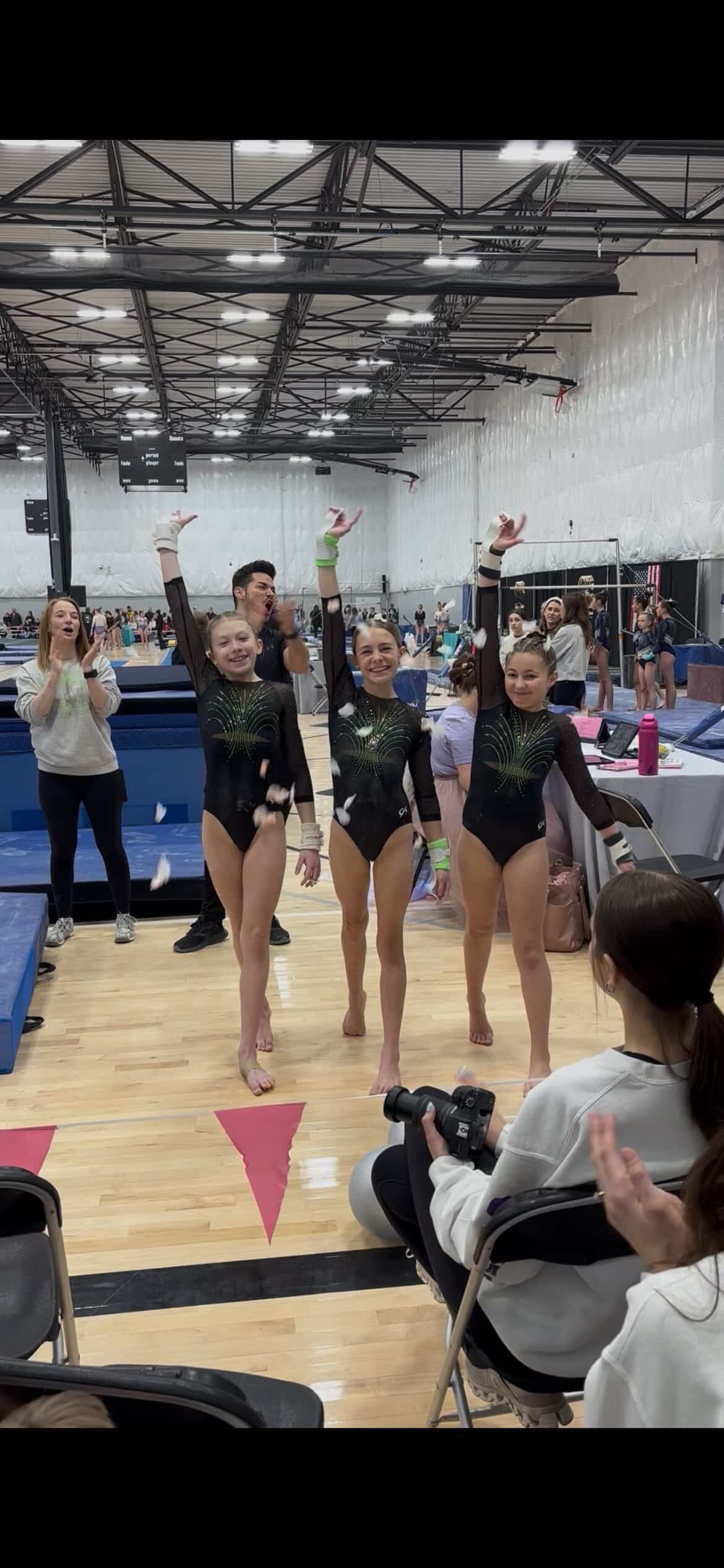 Gymnastics team posing on a gym floor, arms raised in matching leotards during a competition.