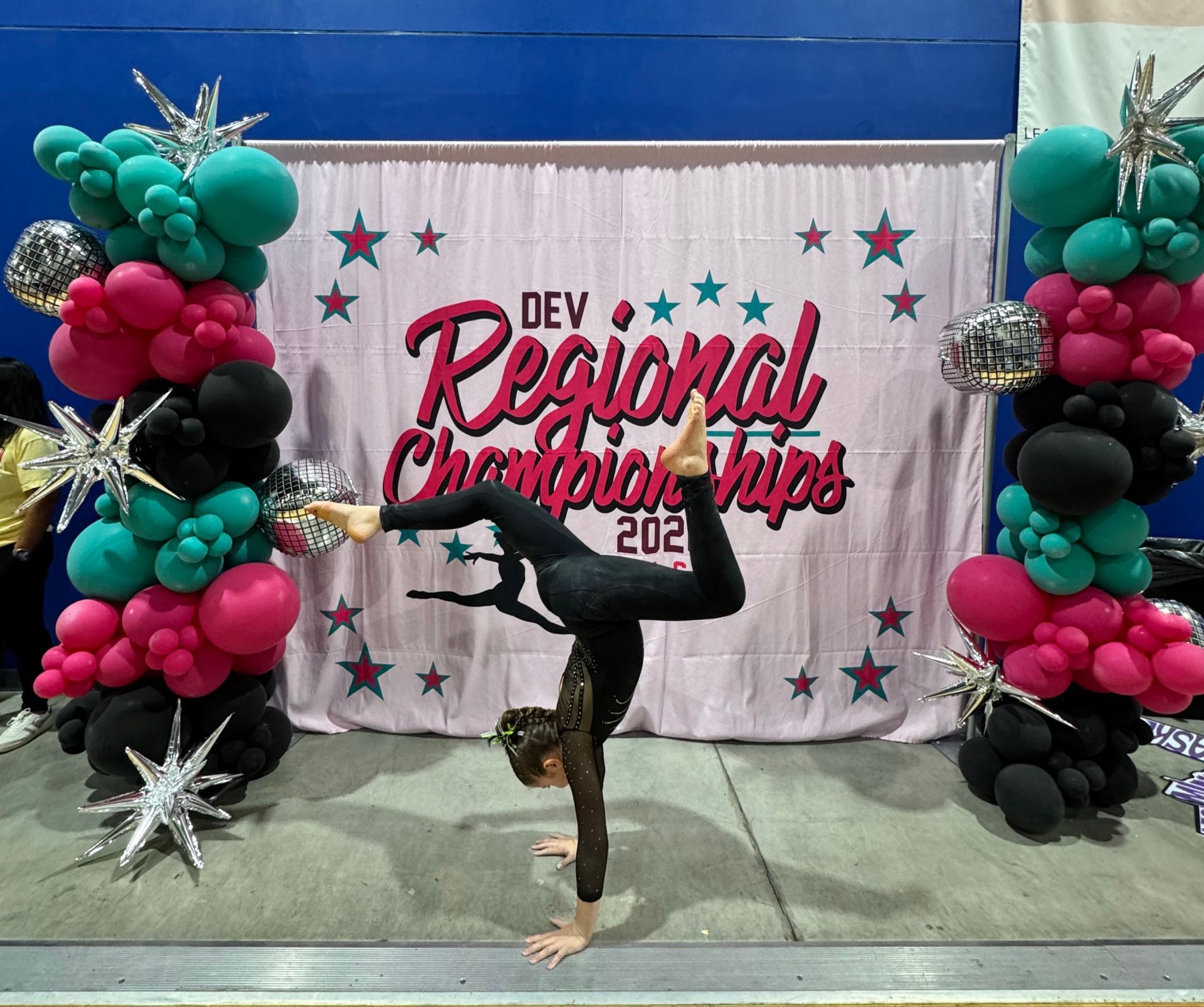 Dancer balancing in a handstand at a Regional Championships backdrop with teal, black, and pink balloon columns