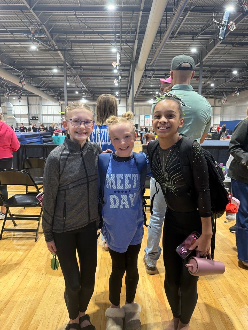 Three people pose smiling in a large indoor venue with folding chairs and bright overhead lights.