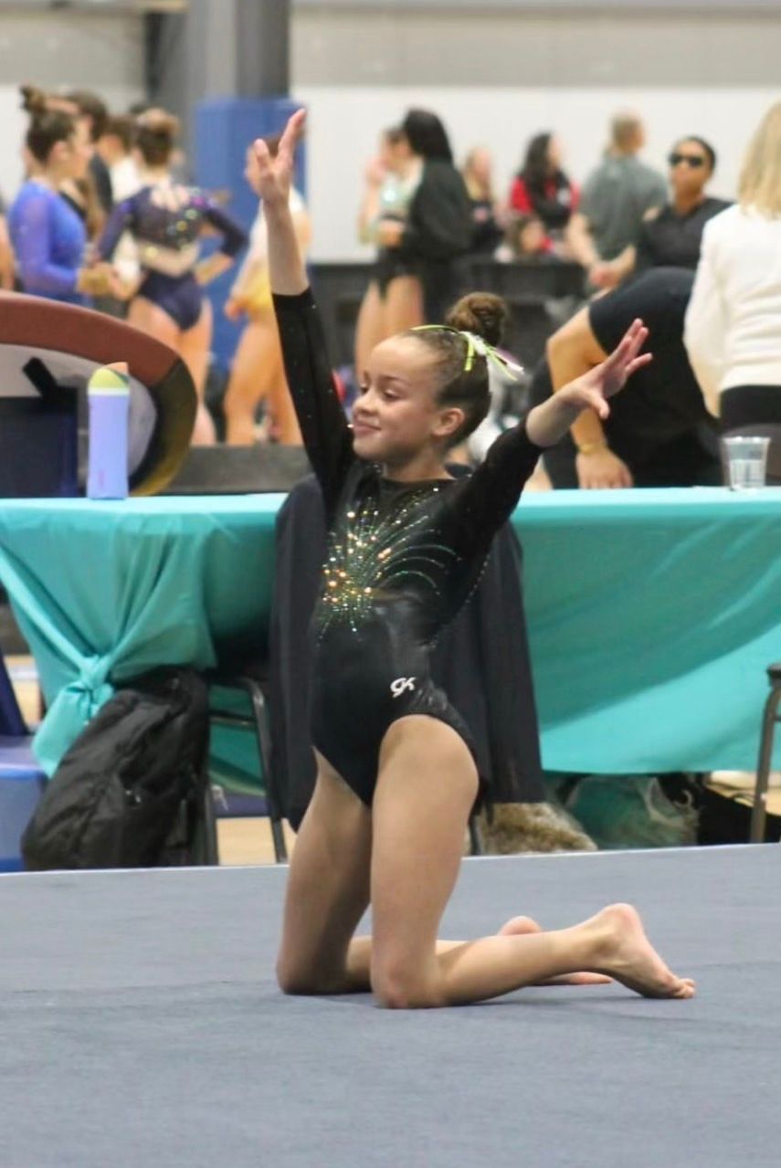Young gymnast in black leotard kneels on a mat with arms raised in a gymnasium competition setting