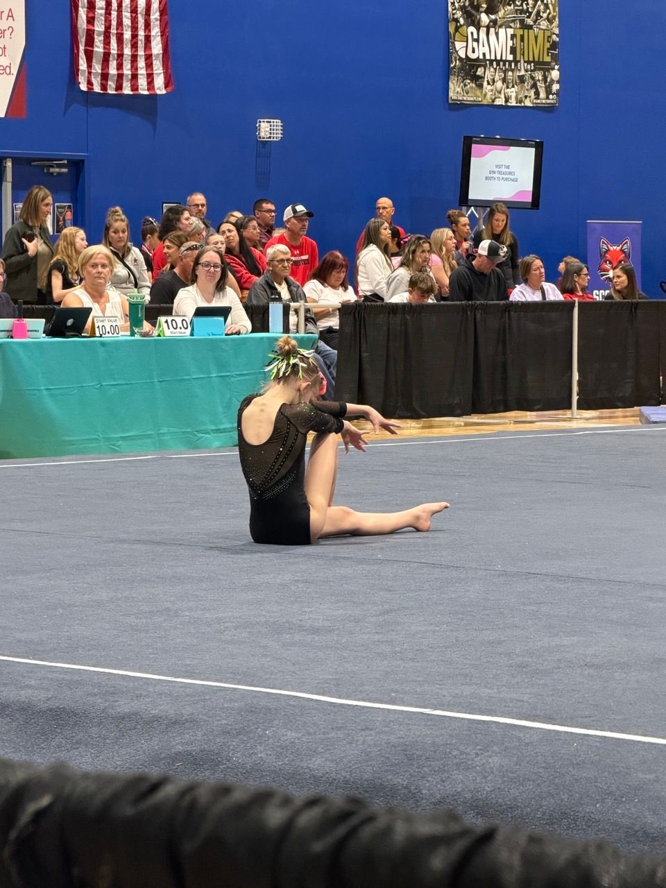 Gymnast seated on a blue mat in a gymnasium, with judges and spectators watching in the background