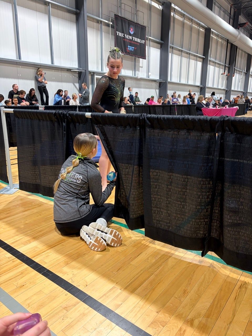 Two people at a black curtain barrier in a gym, one kneeling and handing something through an opening
