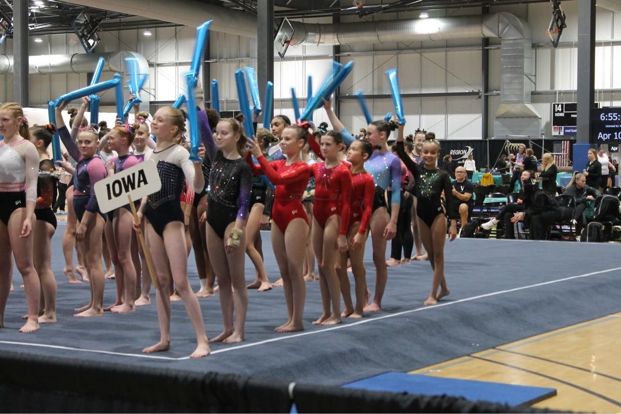Gymnasts lined up on a mat at an indoor meet, with Iowa uniforms visible in front.