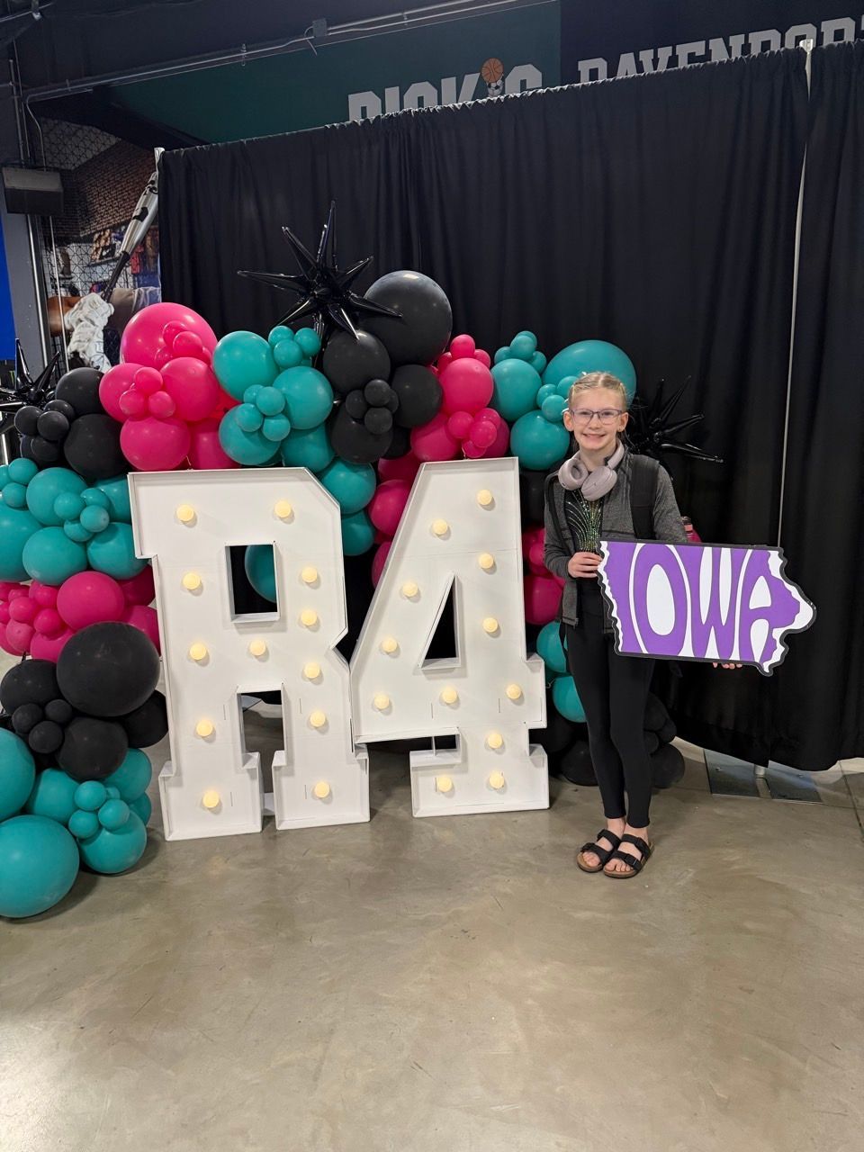 Woman beside lit “84” display holding purple “Iowa” sign with teal, pink, and black balloons in a venue