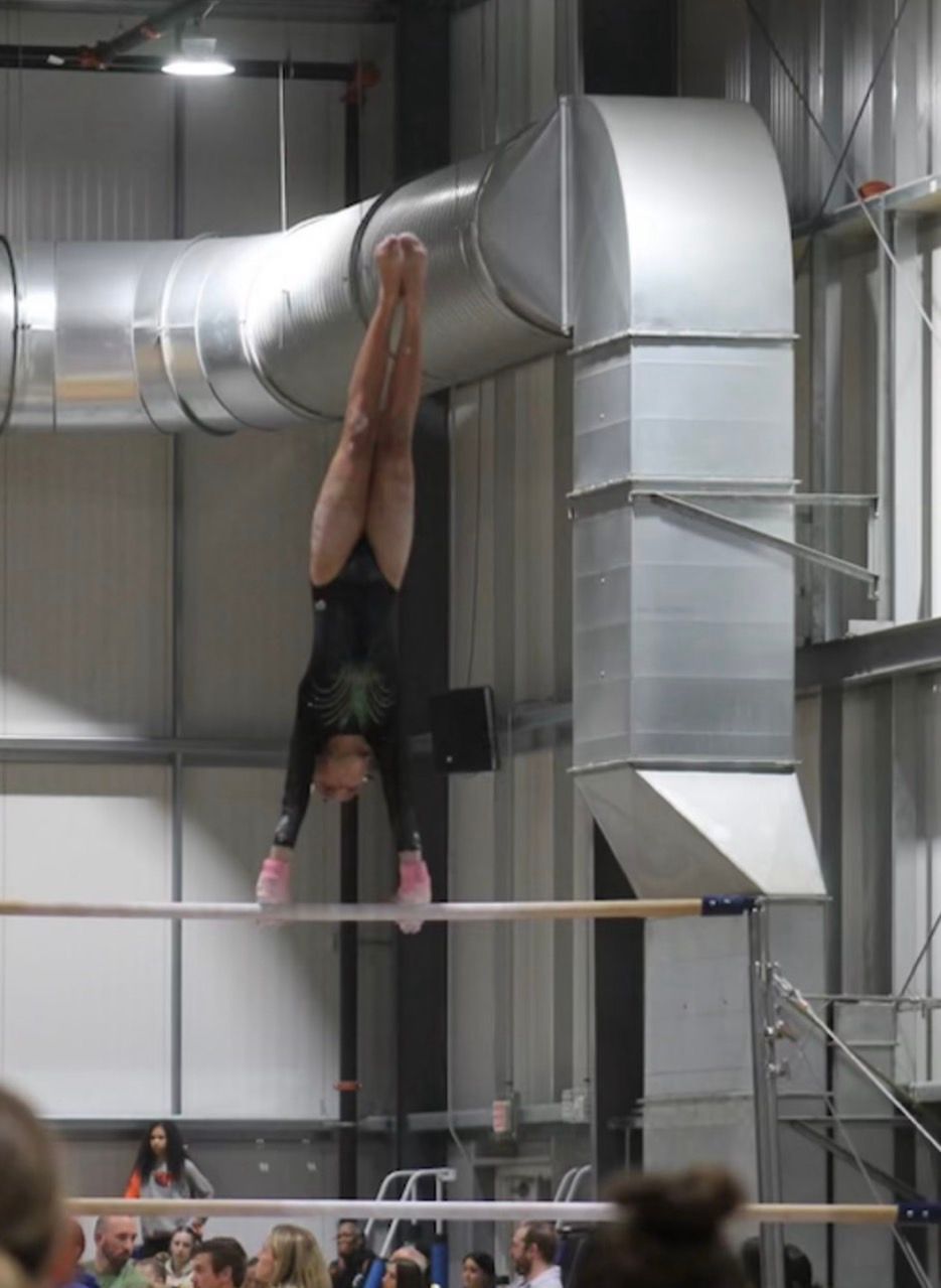 Gymnast hanging upside down on a high bar in an indoor gym, beneath large silver ventilation ducts