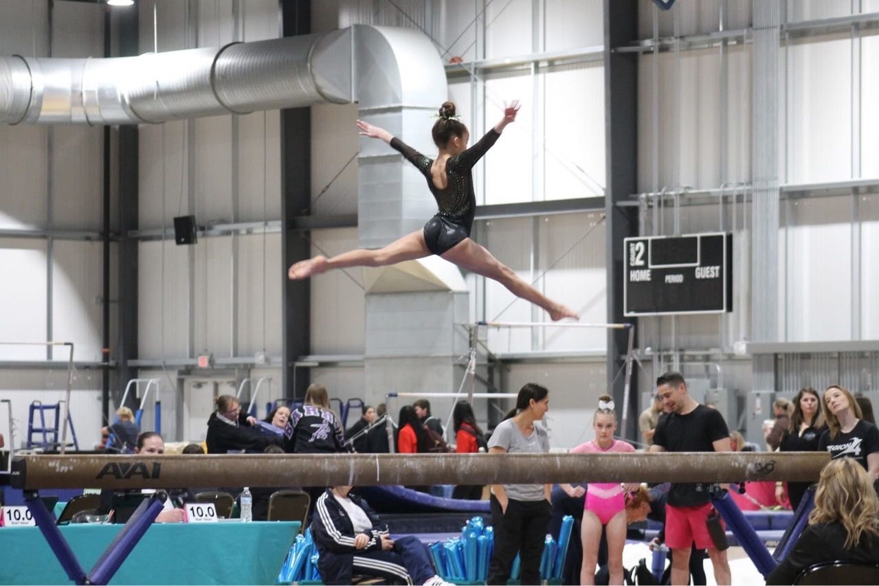 Gymnast midair in a split jump over a balance beam inside an indoor gymnasium