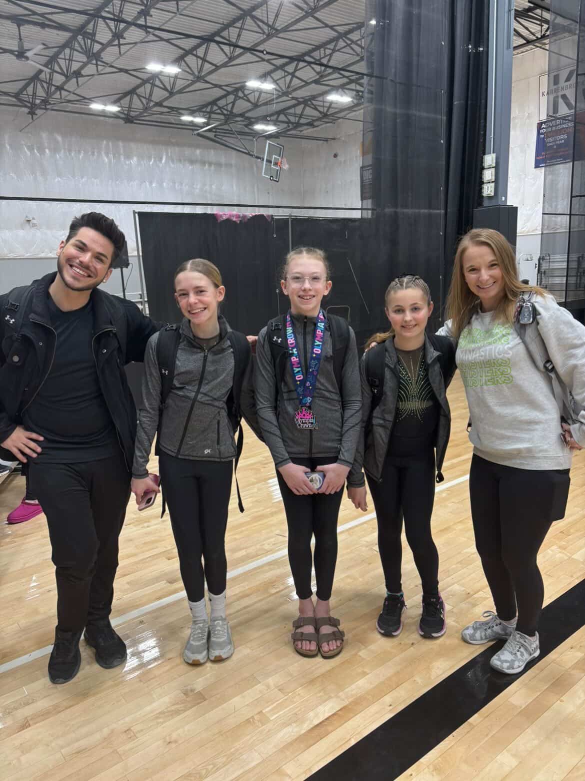 Five people stand together in a gym, smiling on a wooden floor with a black curtain backdrop.