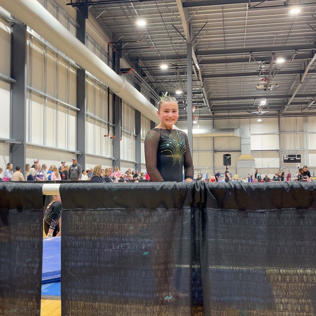Gymnast standing behind a black barrier in a large indoor arena, with spectators in the background