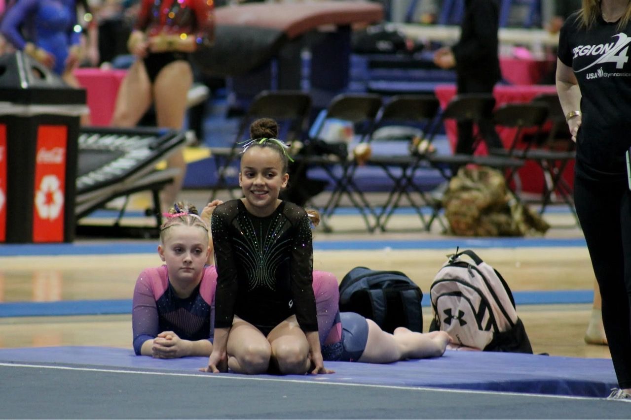 Three gymnasts posing on a mat in a gymnasium, with blurred spectators and equipment in the background.