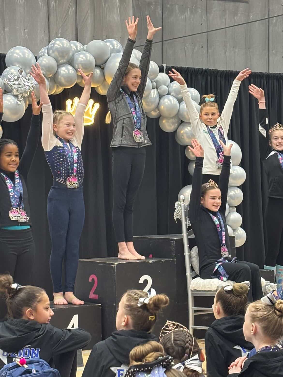 Children on a podium cheering with arms raised during a performance, silver balloons behind them