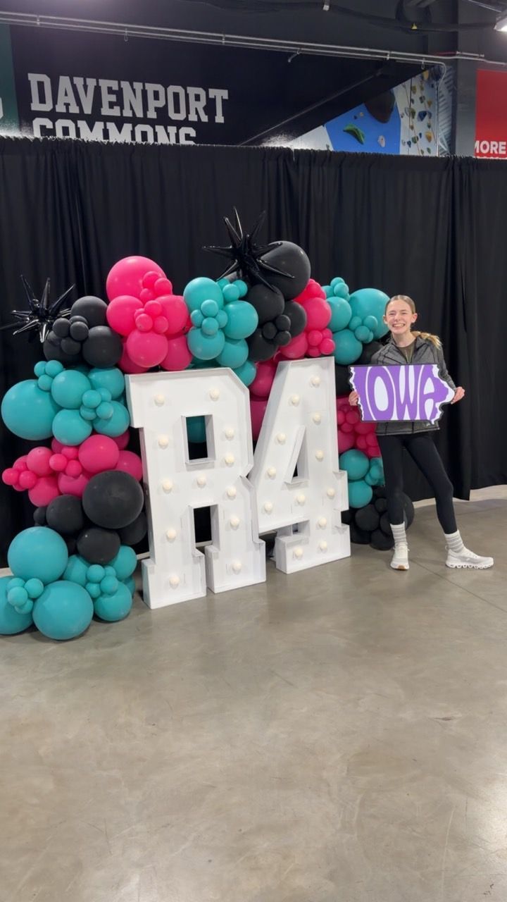 Person posing by large “BA” letters and teal, pink, and black balloons at an indoor event