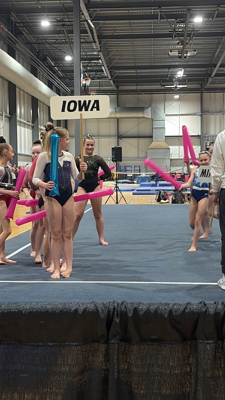 Gymnastics team posing on a blue mat at an indoor meet, with an Iowa sign overhead.