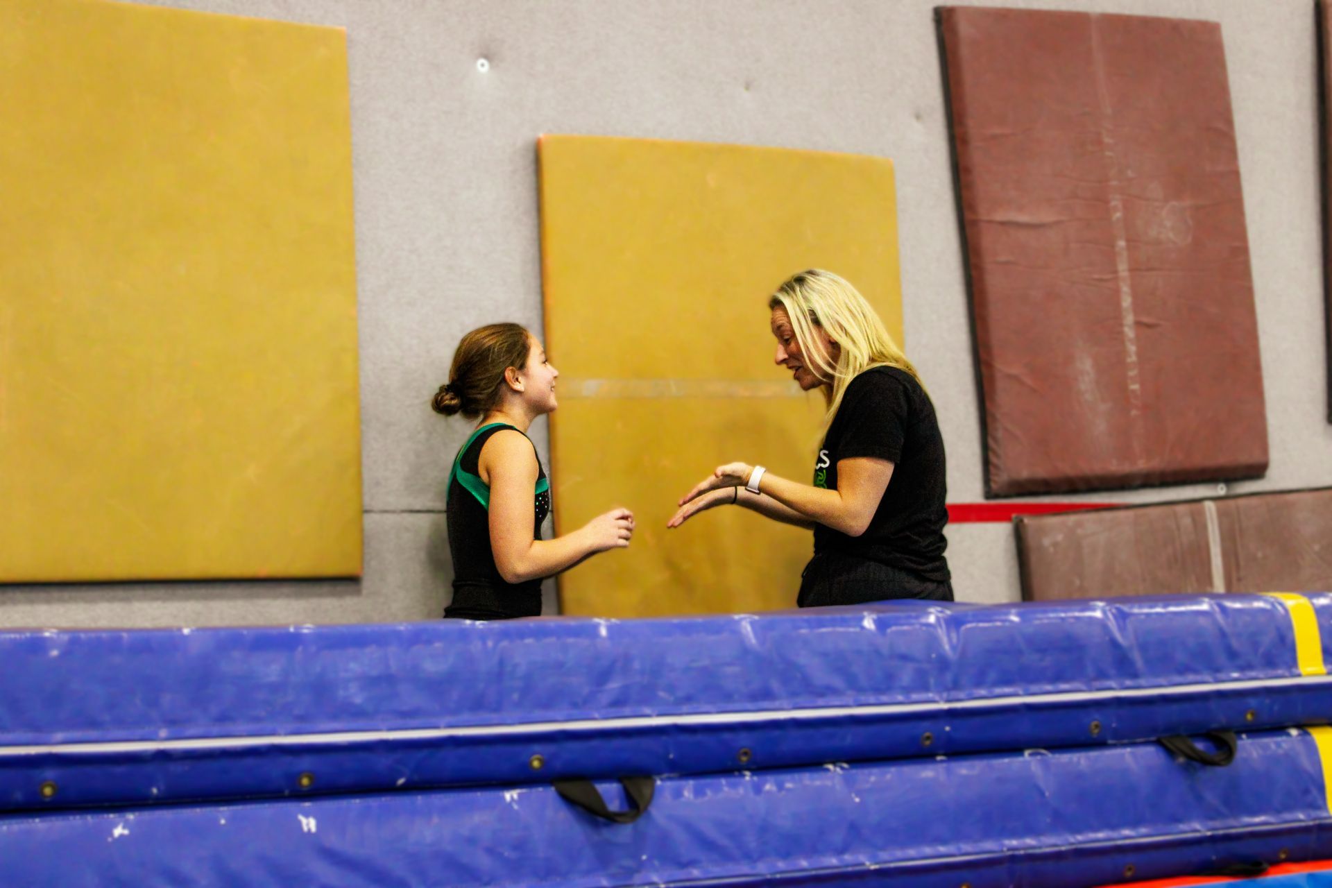 Two people talk beside a blue mat in a gym with yellow wall pads.