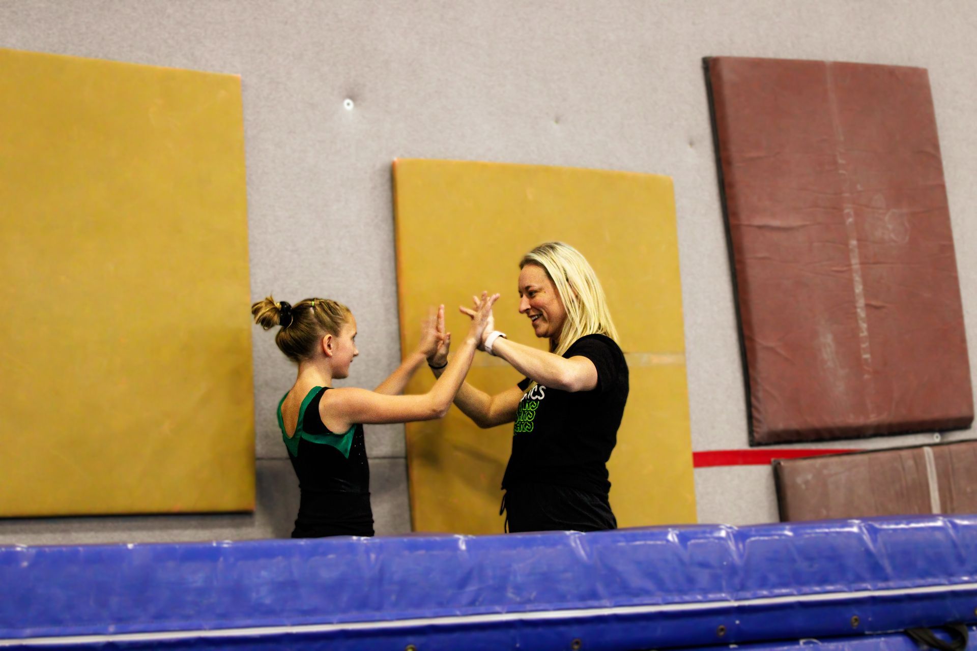 Two people in a gym spot each other with raised hands near foam mats.