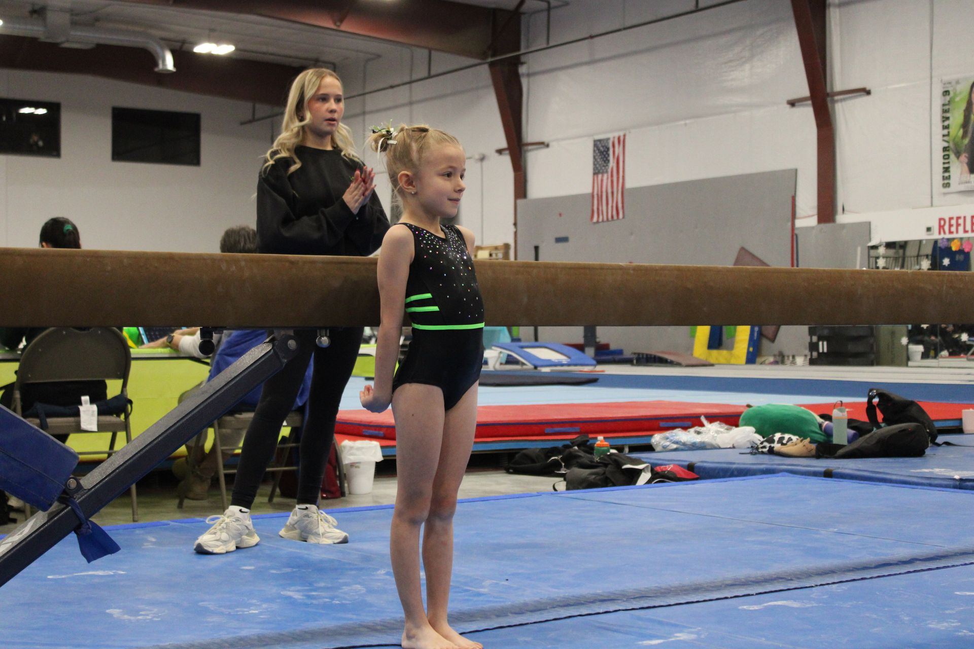 Young gymnast standing beside a balance beam in a gym, with a coach behind her