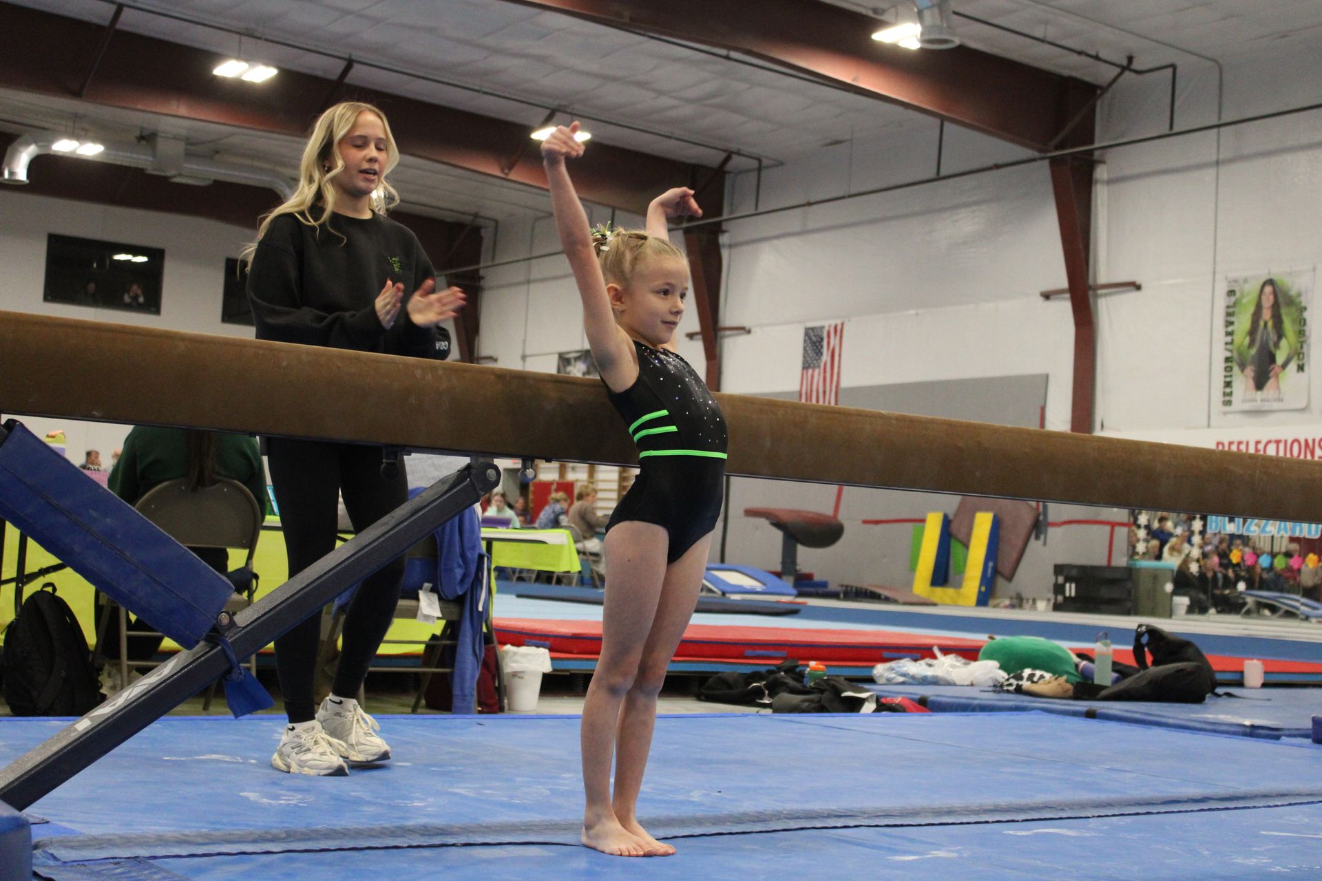 Gymnast in black leotard raises arms beside a coach in a gymnastics gym.