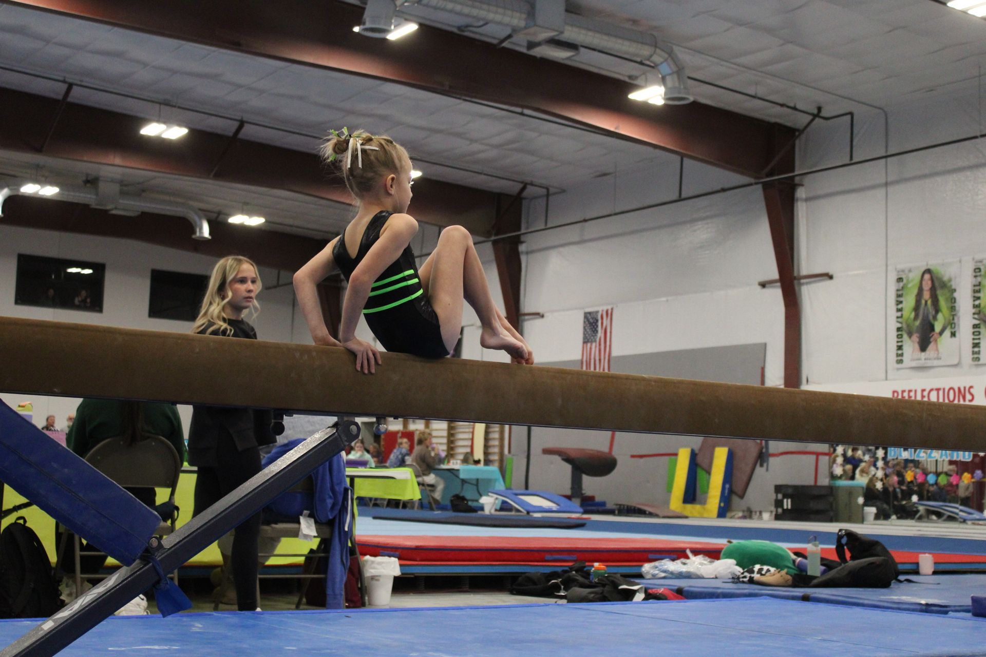 Gymnast balancing on a beam in a gymnastics gym, with mats and equipment in the background.