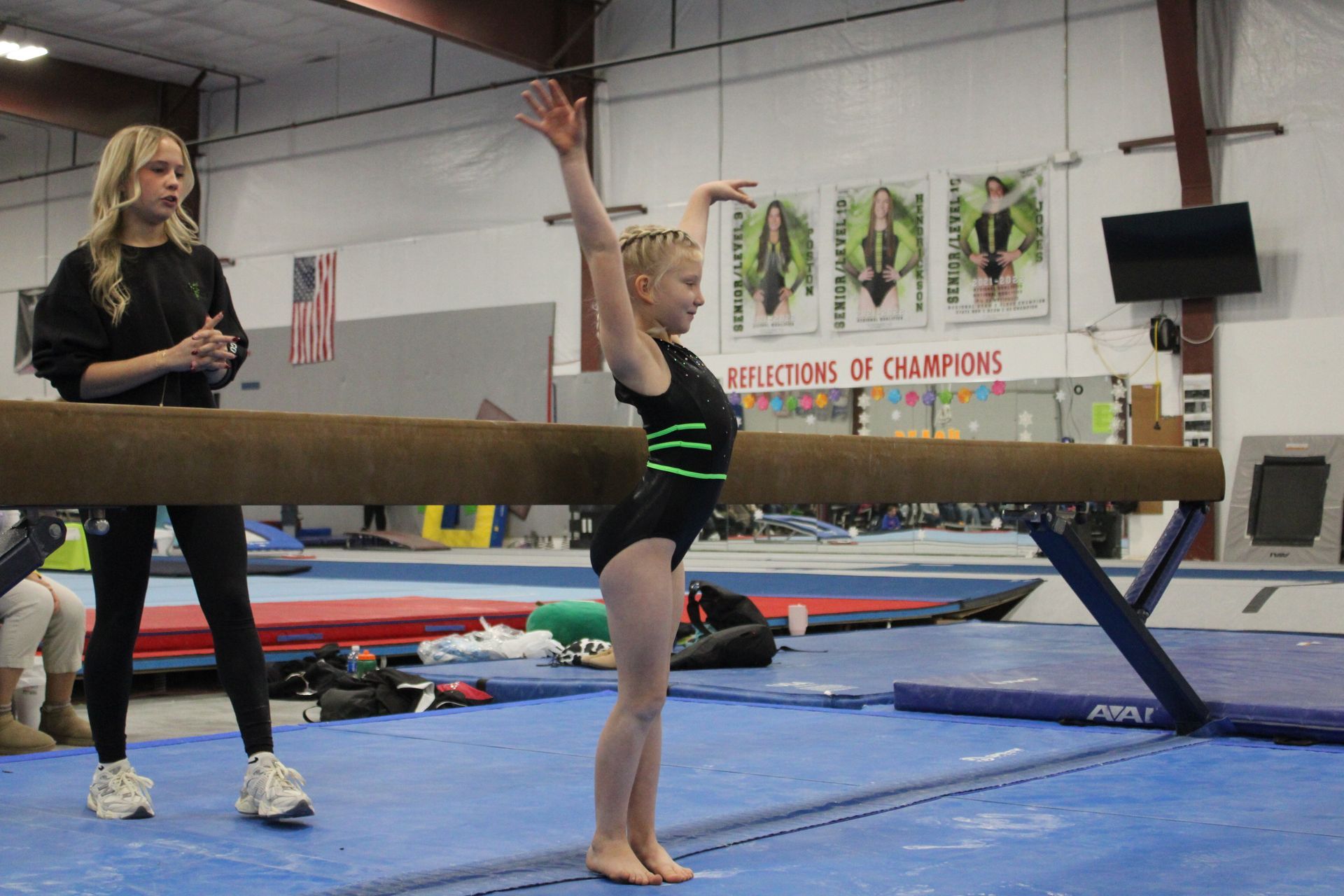 Gymnast balancing on a beam in a gym, with a coach watching nearby.