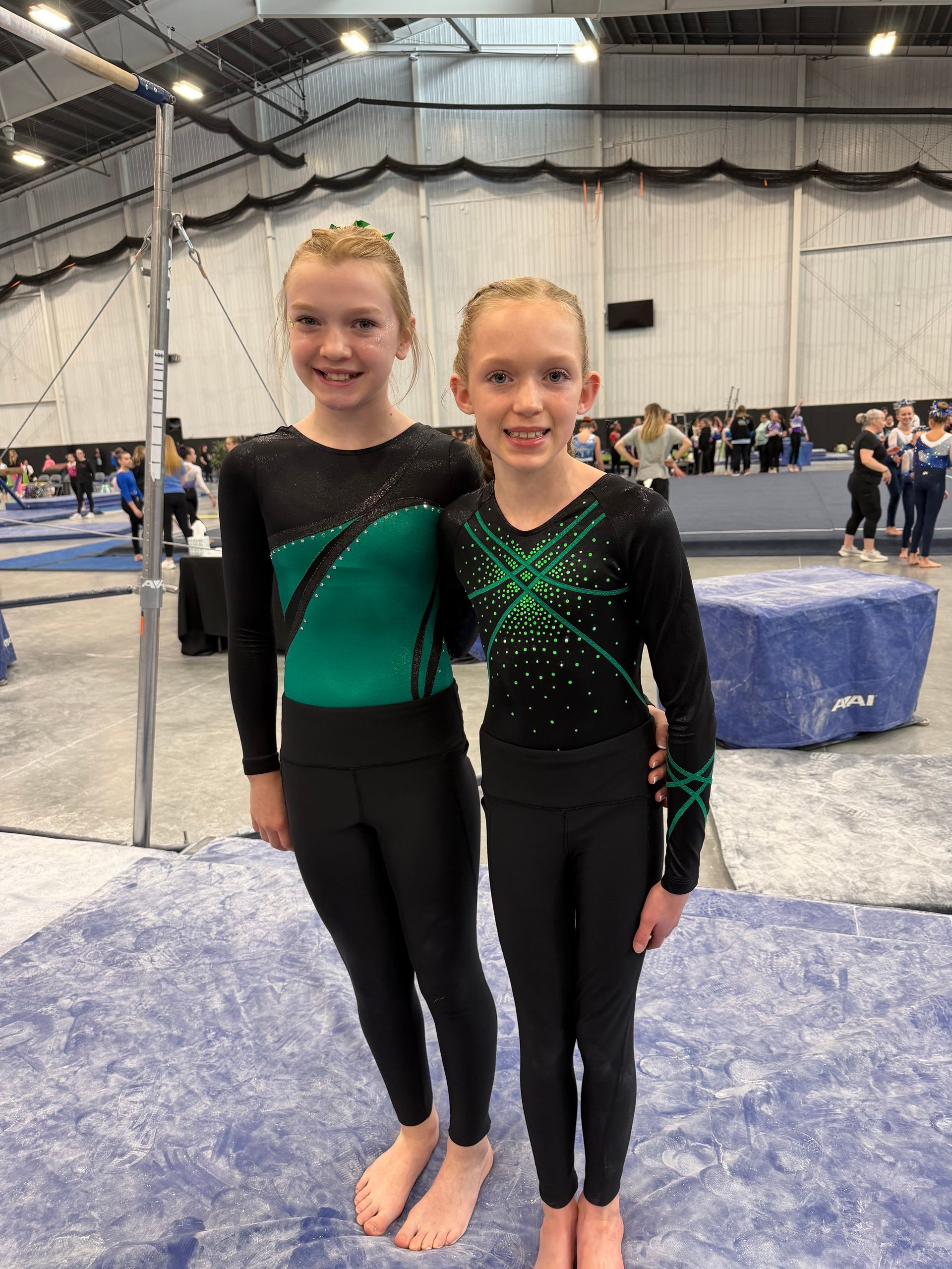 Two gymnasts in matching black and green leotards standing together at an indoor gymnastics meet.