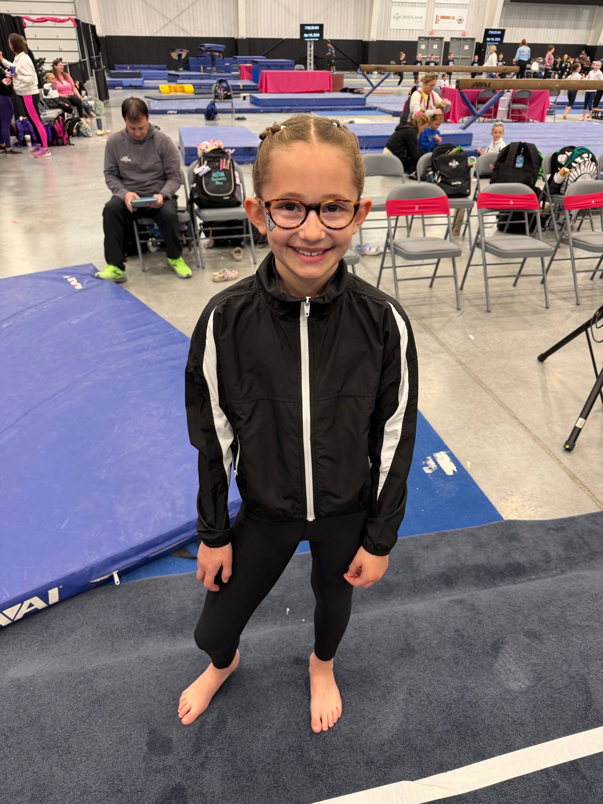 Smiling child in black jacket and glasses standing barefoot on a gym mat at an indoor event
