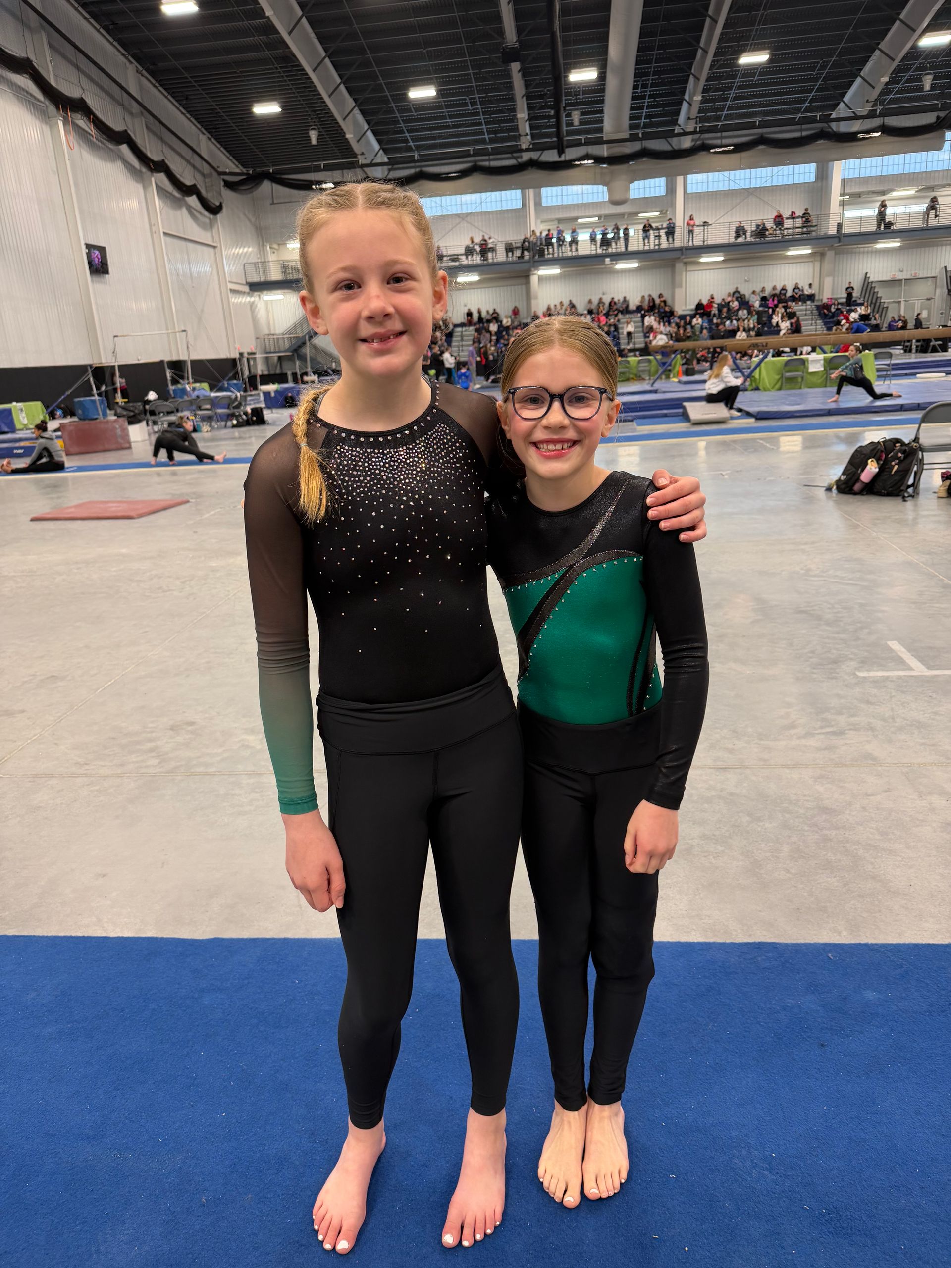 Two young gymnasts smiling in black leotards on a blue mat inside a large indoor arena.
