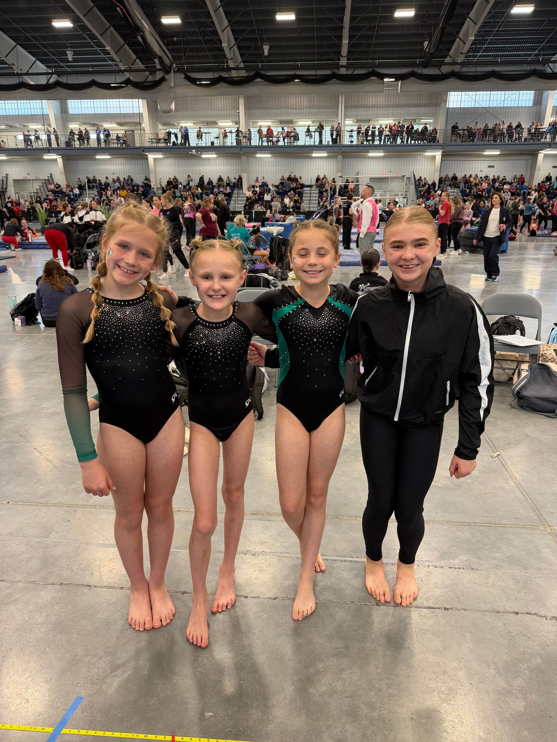 Four young gymnasts in black leotards pose on a competition floor inside a crowded arena.