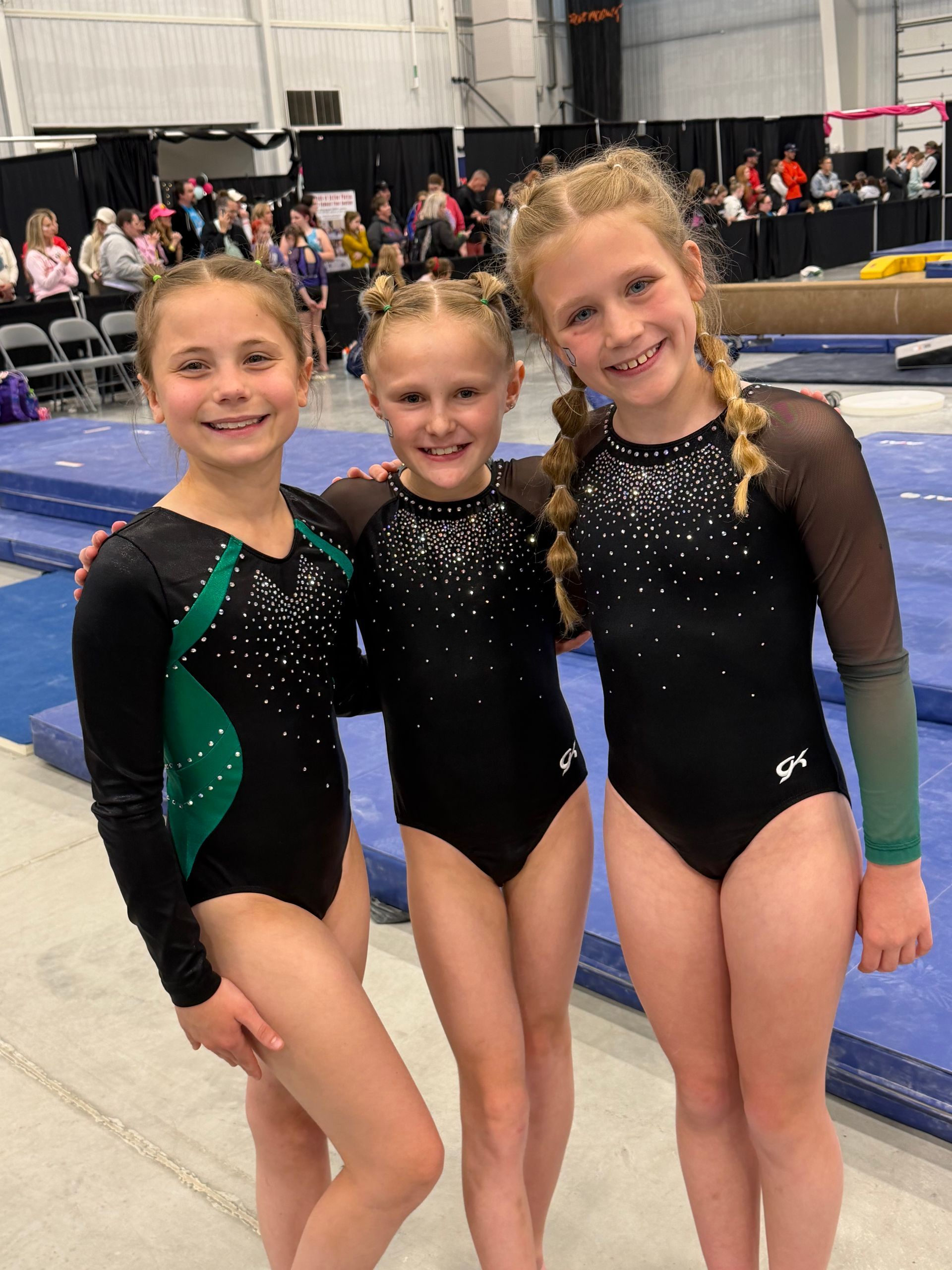 Three gymnasts in black leotards pose smiling at a gymnastics arena.