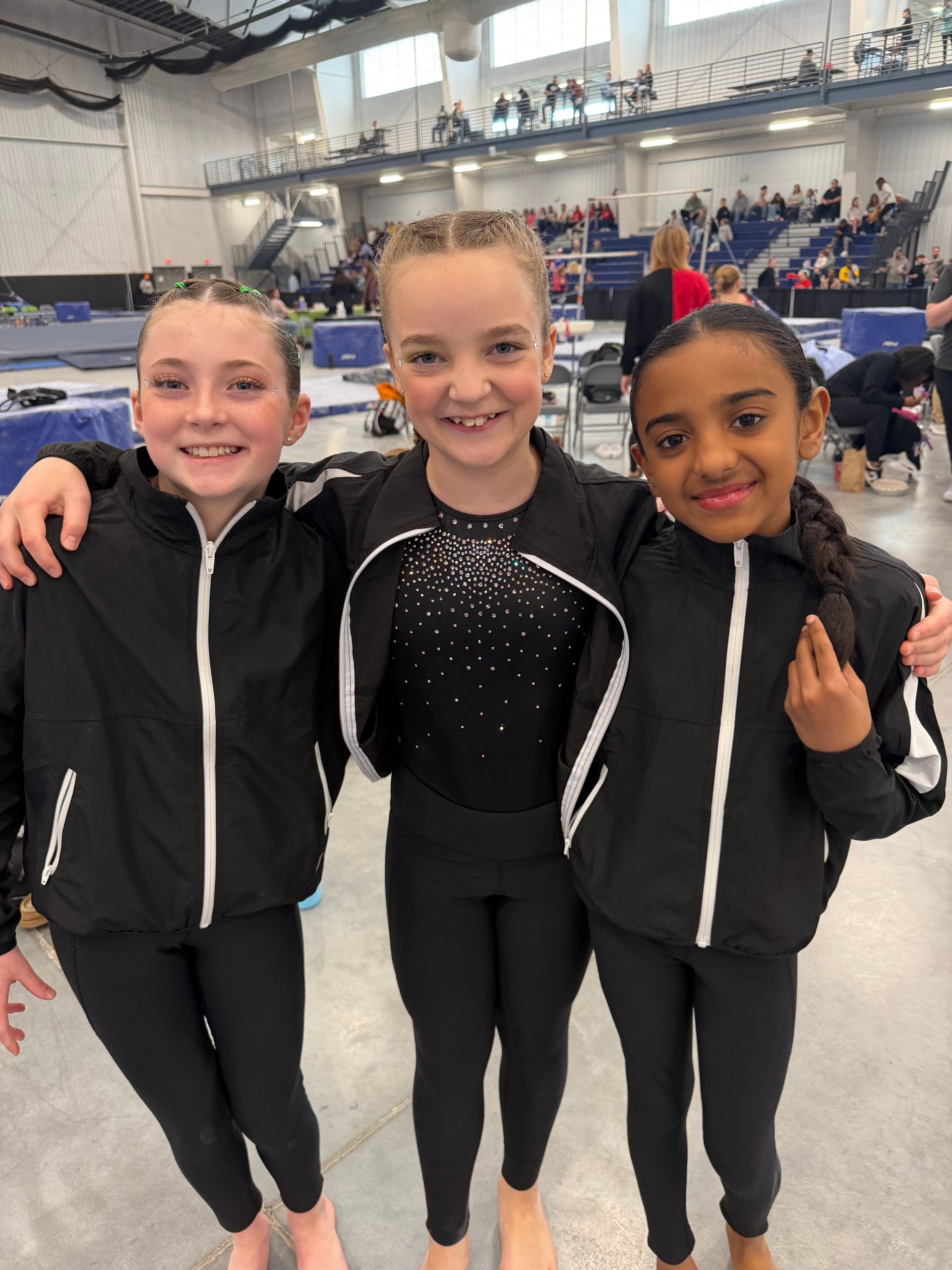 Three gymnasts in black leotards smiling together in an indoor arena