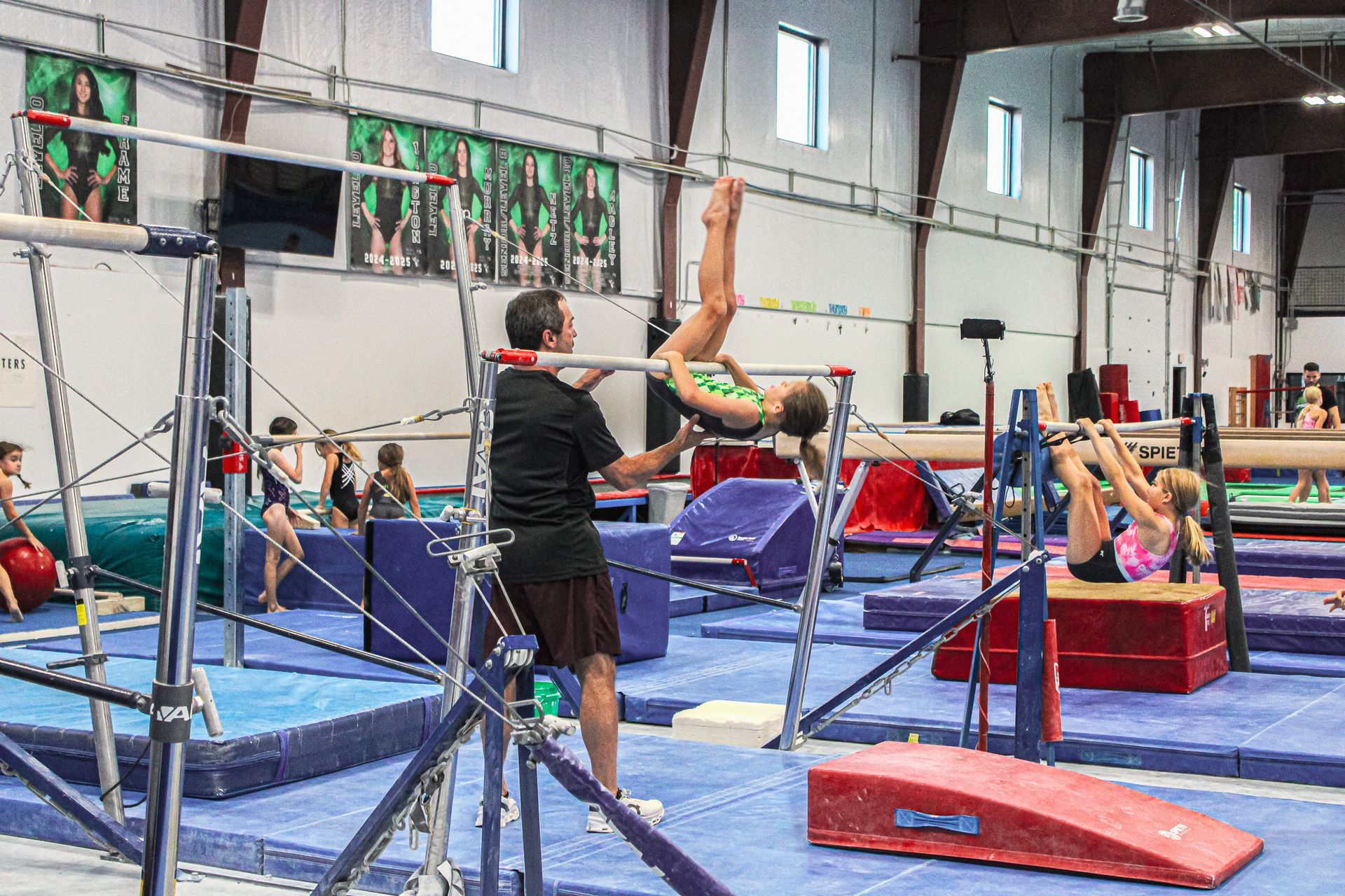 Gymnastics training in a gym, with a coach spotting athletes on uneven bars and mats.