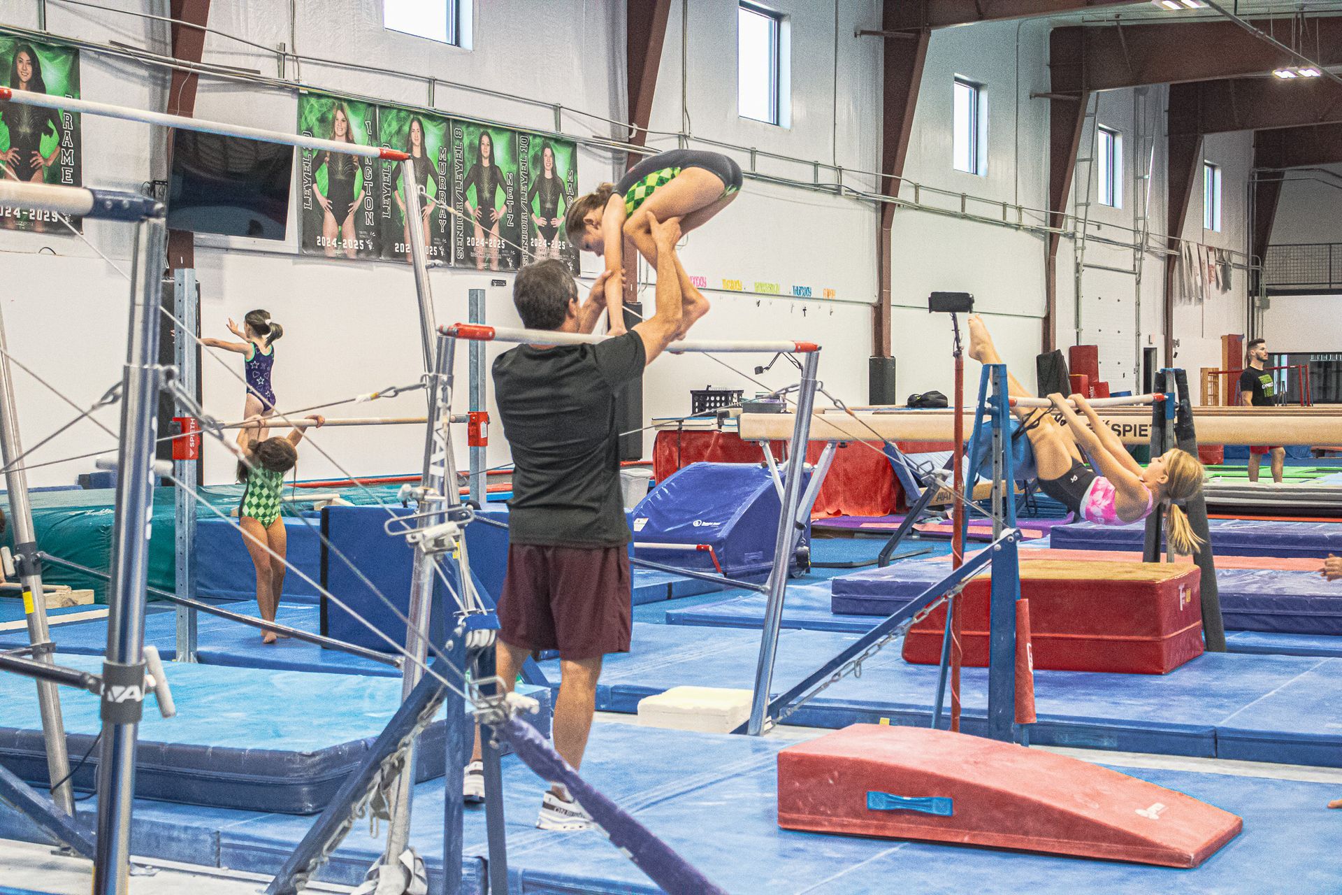 Gymnastics practice in an indoor gym, with a coach spotting a gymnast on the uneven bars.