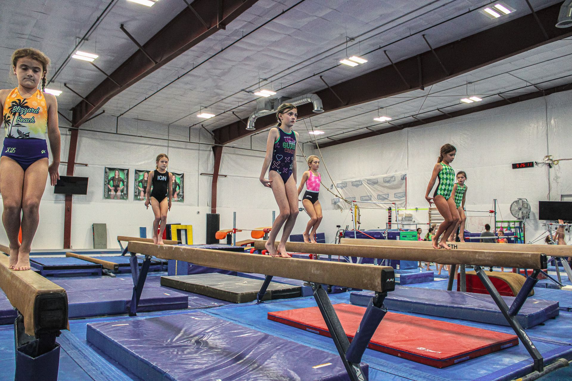 Gymnasts practicing balance beam routines in an indoor gym with mats and trampolines