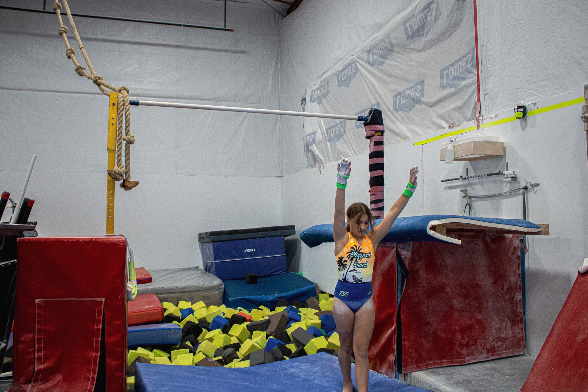 Gymnast standing with arms raised in a foam pit area, with red mats and gym equipment around her.