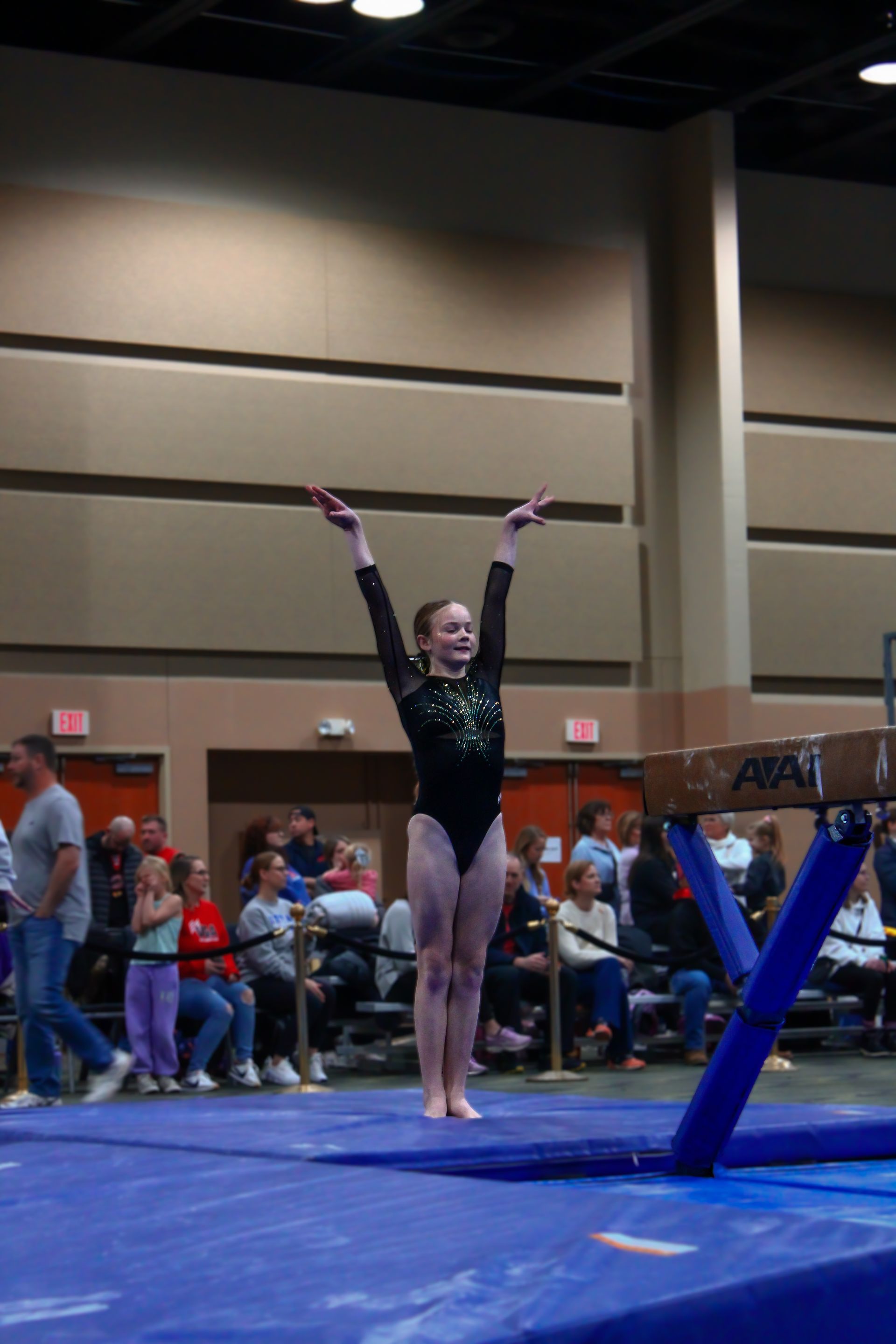 Gymnast in black leotard posing with arms raised on blue floor mat at an indoor meet