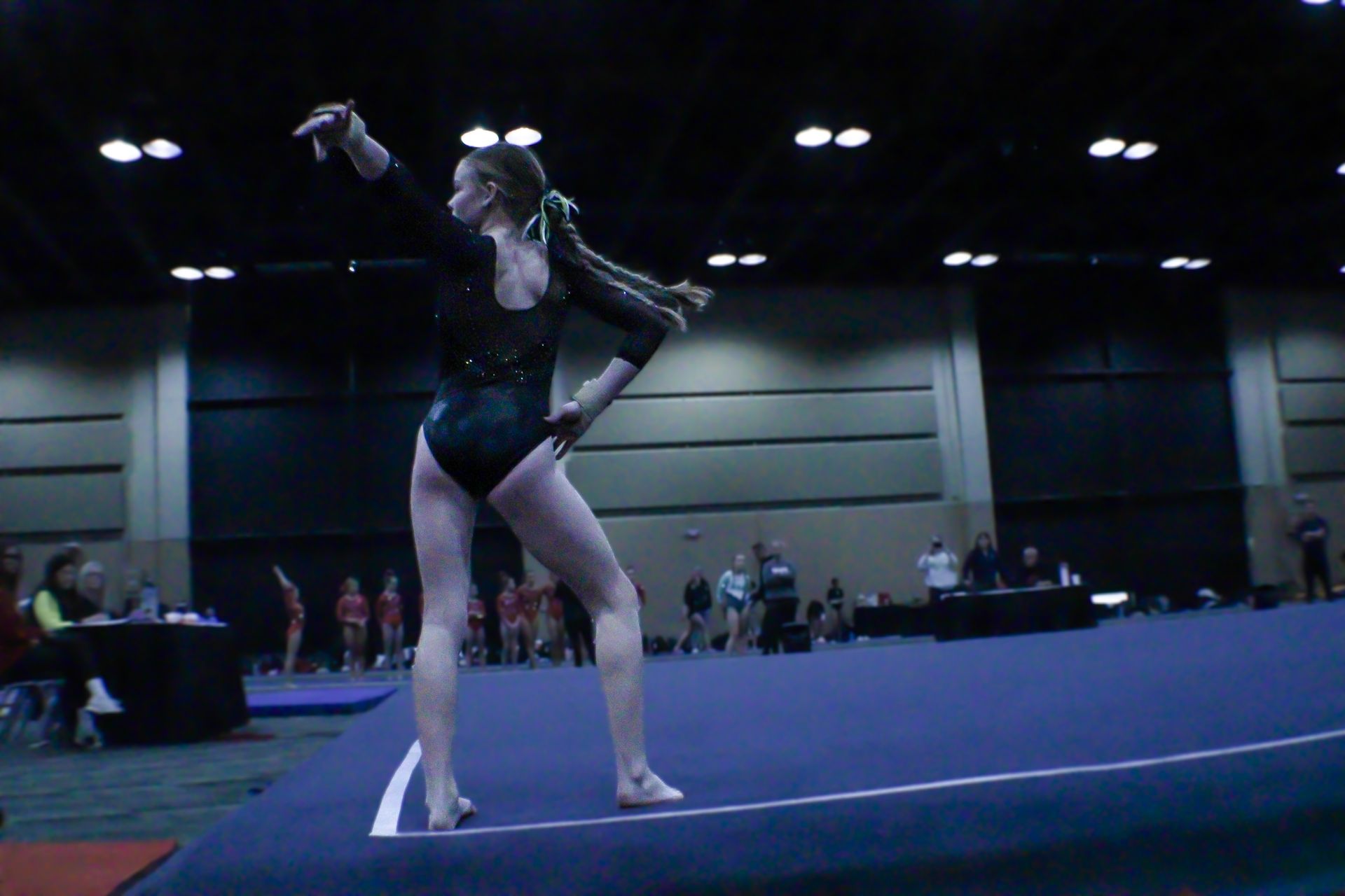 Gymnast posing on a blue mat in a competition hall, wearing a black leotard.