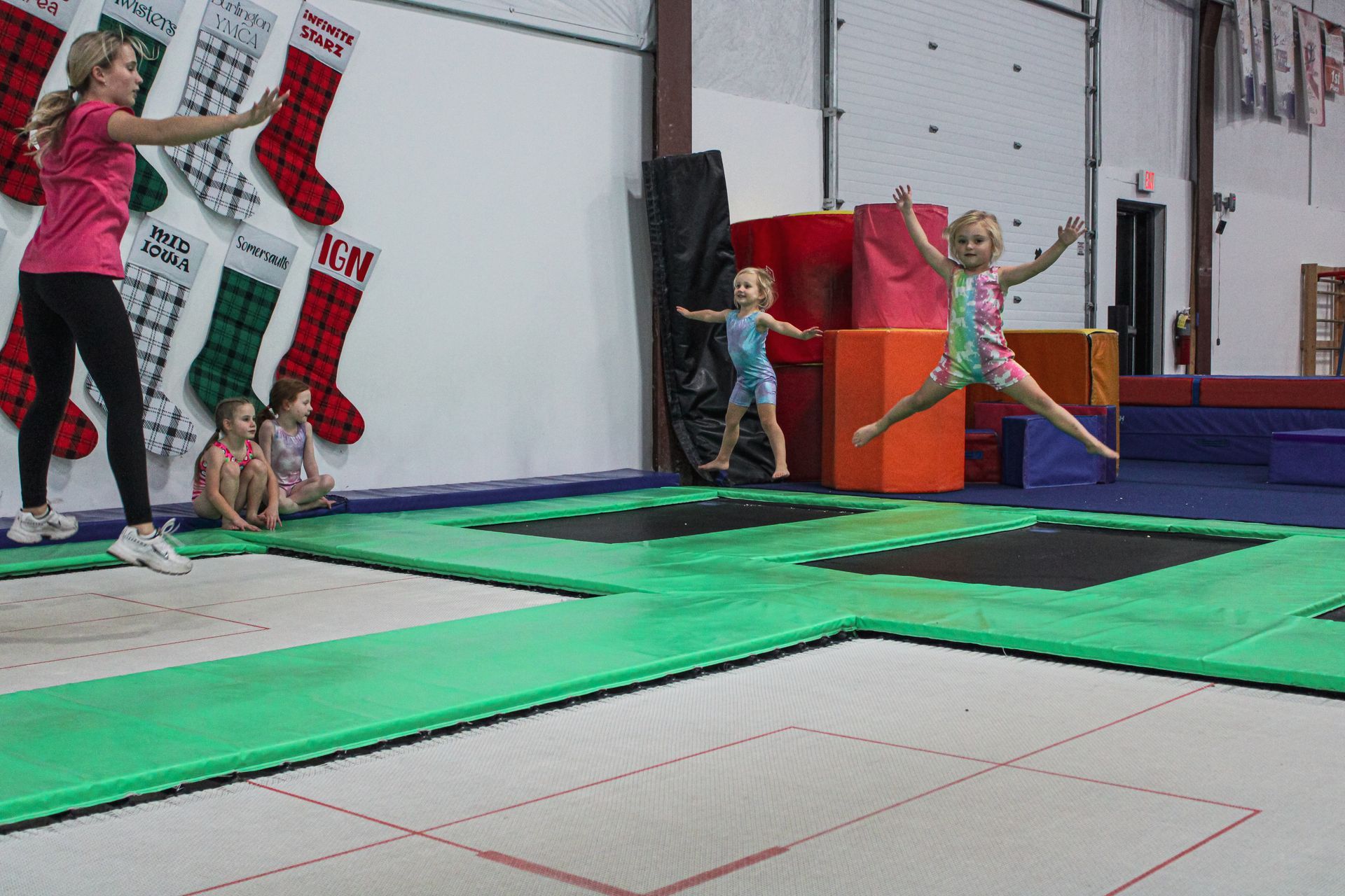 Children bouncing and jumping on green trampolines in an indoor gym with holiday stockings on the wall