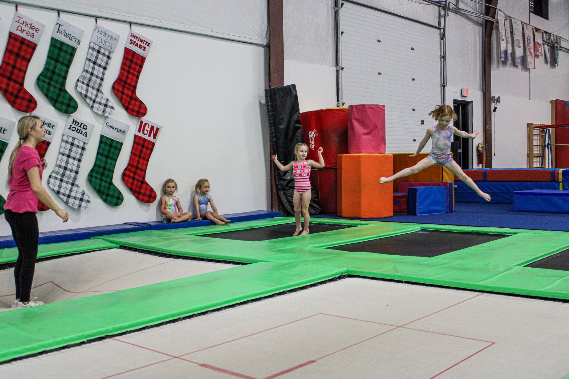 Children on colorful trampolines in a gymnastics gym, one mid-jump and others waiting nearby